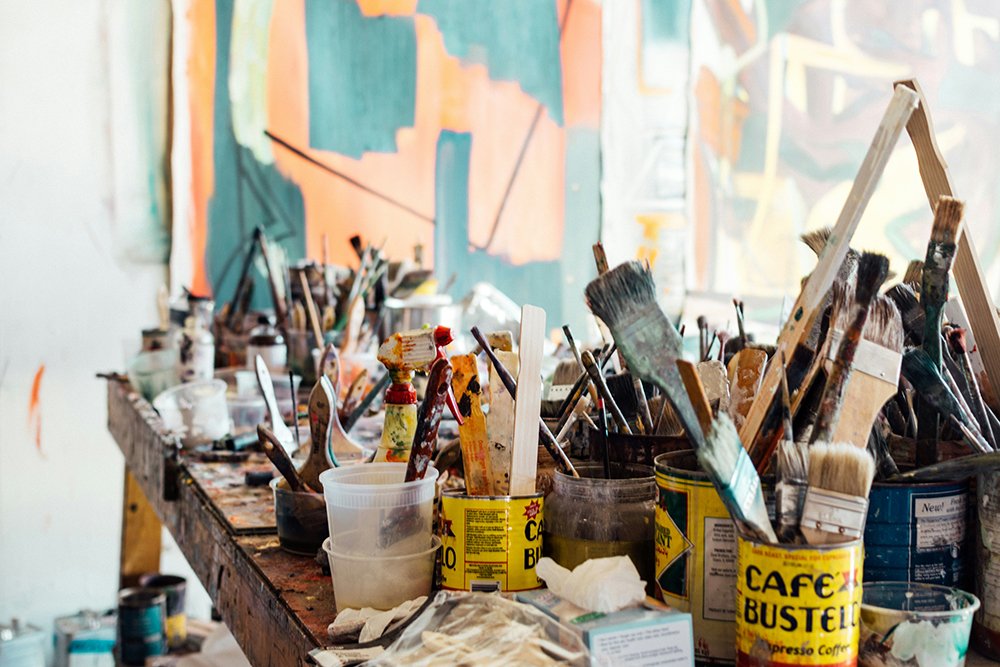 Paint brushes, paint and other artist tools scattered on a benchtop in a studio