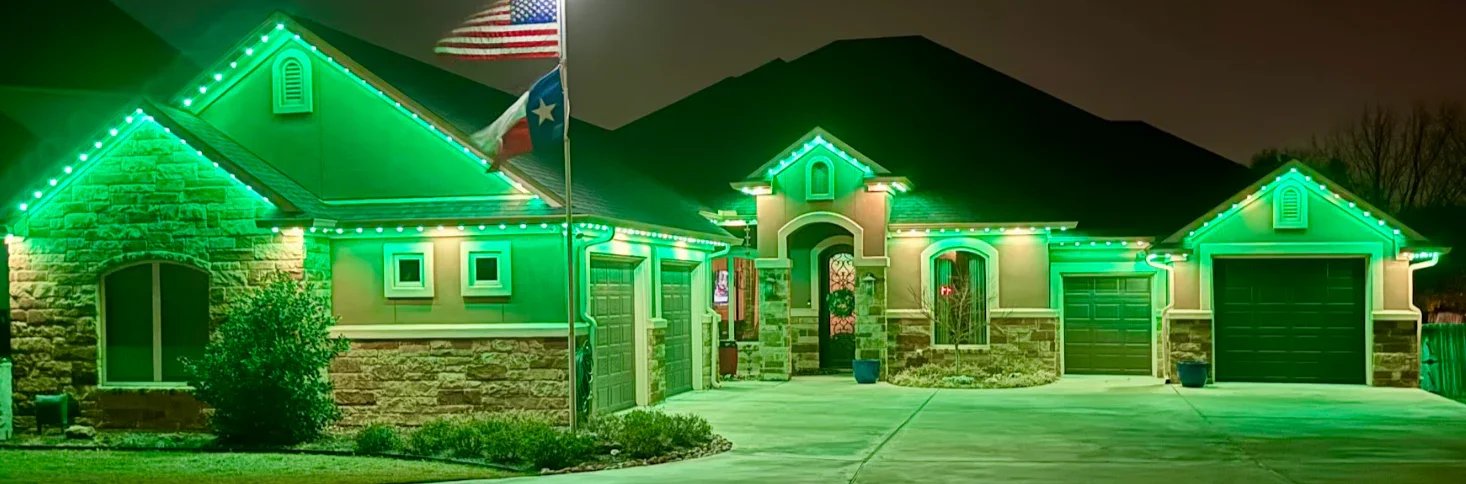 A house decorated with green holiday lights, with an American flag and a Texas flag flying outside, at night.