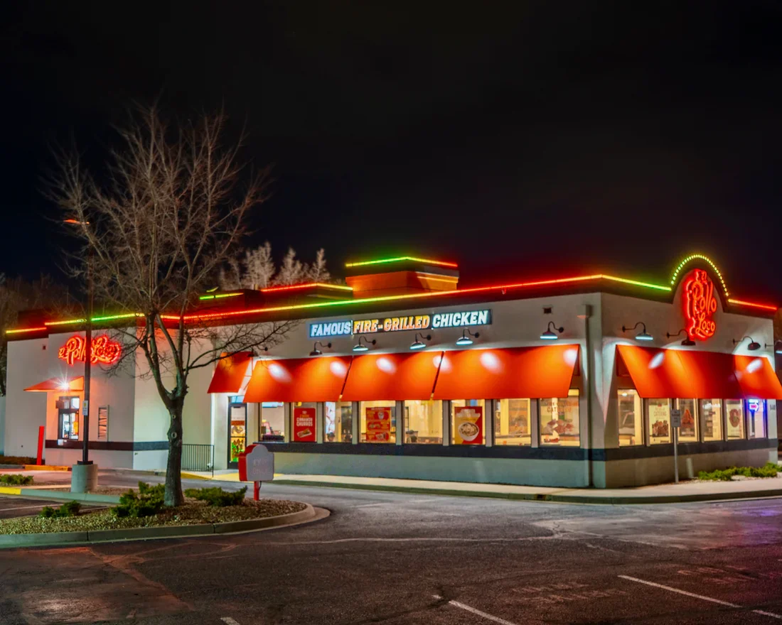 Night view of a fast food restaurant with neon lights, sign advertising fire-grilled chicken, orange awnings, and a parking lot in front.