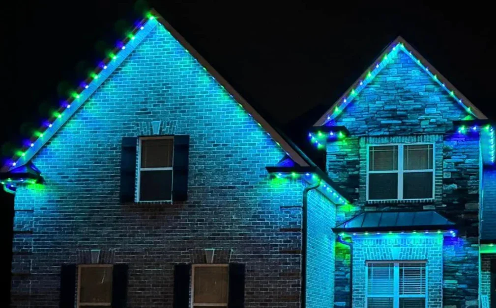 Nighttime view of two houses decorated with blue and green Christmas lights along the rooflines.