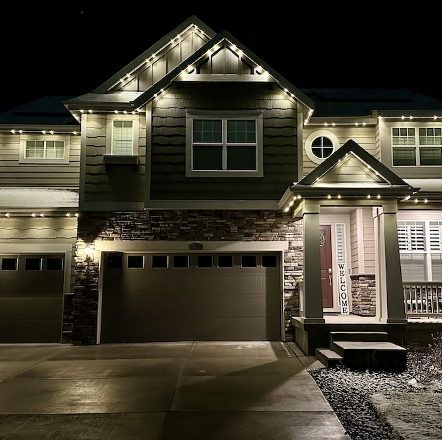 A two-story house at night decorated with Christmas lights, featuring a garage, a front porch with a welcome sign, and illuminated windows.