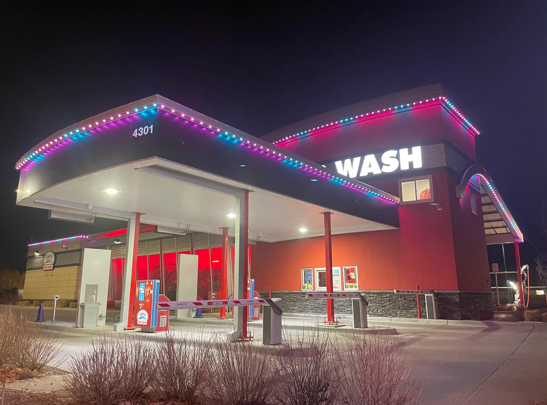 Night view of a brightly lit car wash with purple and pink LED lights, a large sign that reads 'WASH,' and an outdoor payment kiosk at the entrance.