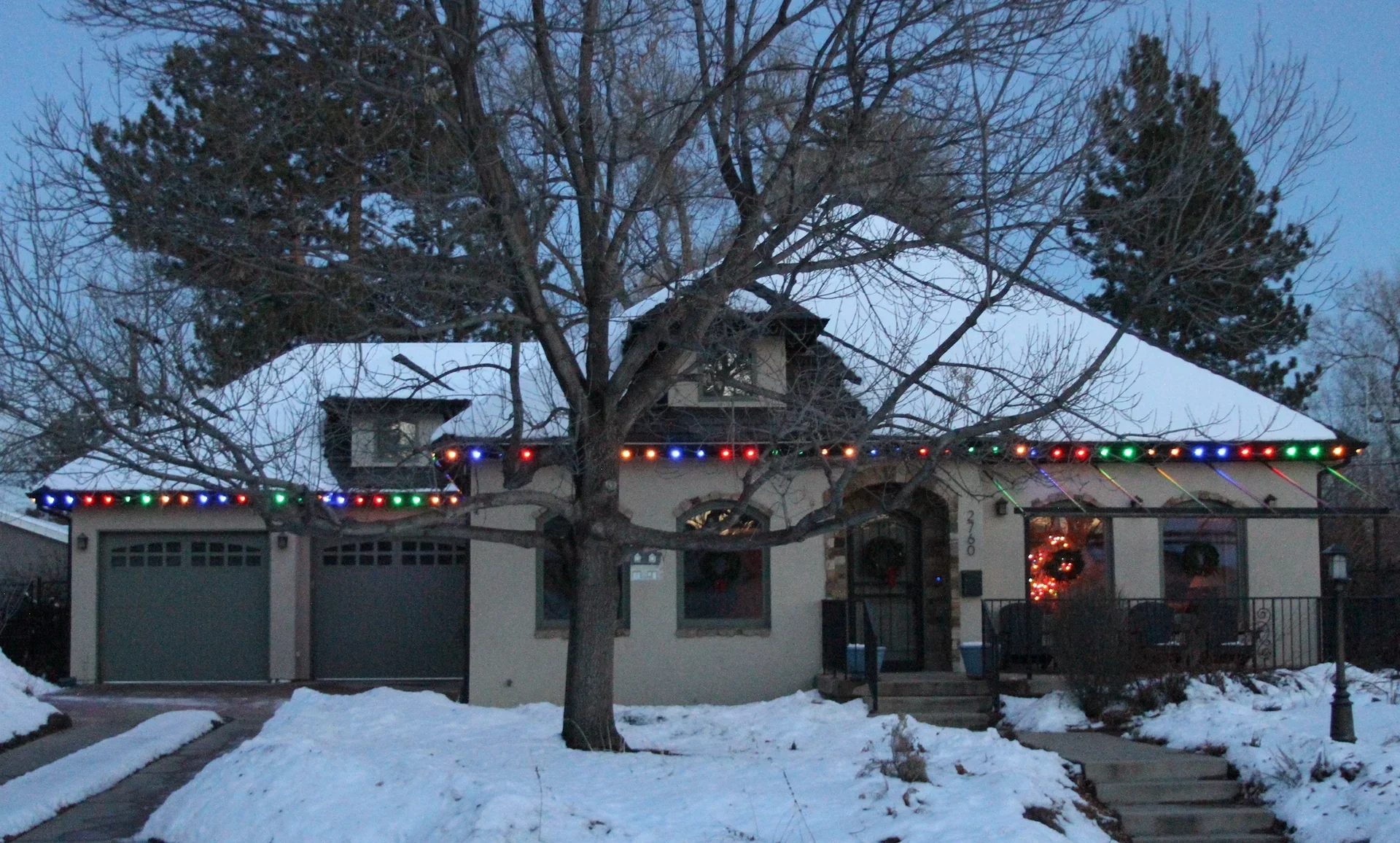 A house decorated for Christmas with multicolored string lights on the roof, a wreath on the front door, and snow on the ground.