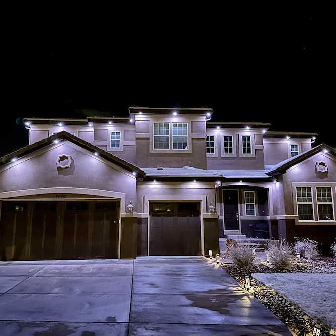 A modern two-story house illuminated with exterior lights at night, featuring a two-car garage, a front porch, and landscaped front yard with plants and rocks.