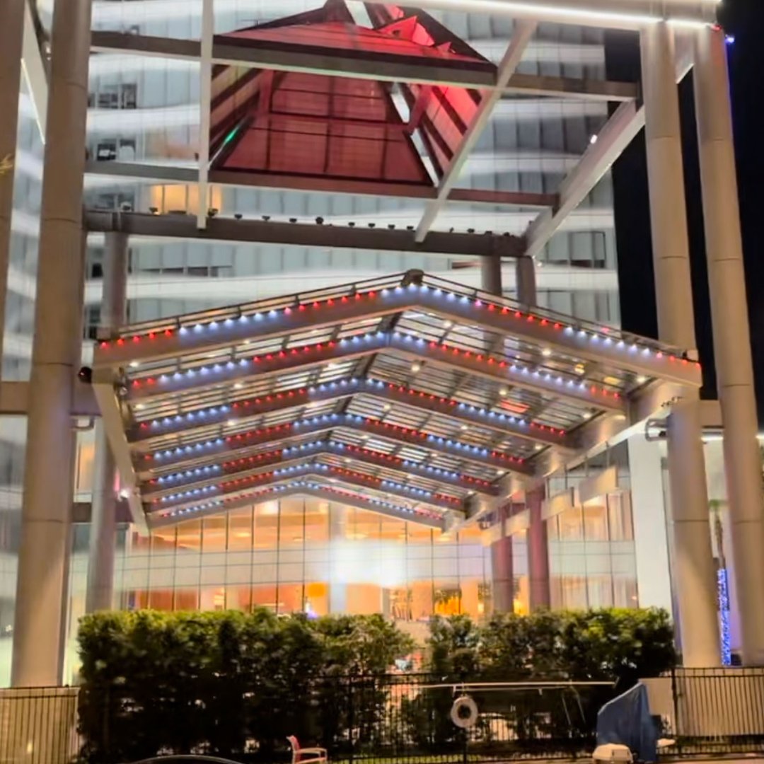 Night view of an outdoor area with a decorated glass and metal canopy illuminated with red, white, and blue string lights, attached to a modern building with large glass windows.