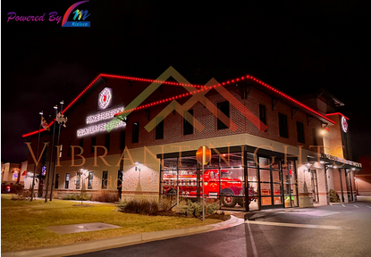 Night view of a two-story building with a glass facade on the ground floor, red lighting accents along the roofline, and a red fire truck parked inside visible through the windows. Street lights and a grassy area with a sidewalk are also in the scene