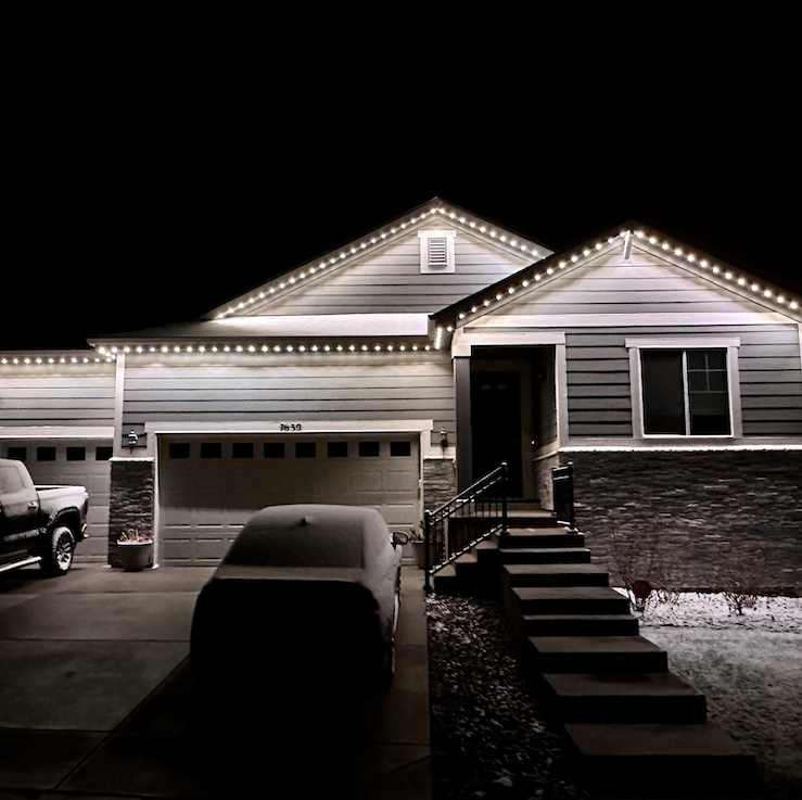 A house decorated with white Christmas lights on the roof at night, with a driveway, a vehicle, and snow on the ground.