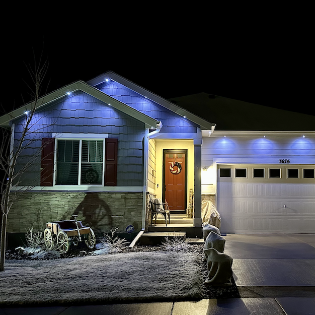 A house decorated with Christmas lights, a wreath on the front door, and a chair on the porch. There is a snow-covered lawn with decorations including a small wagon and a dog statue.