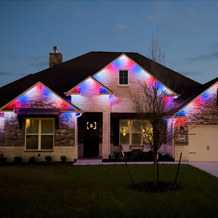 A two-story house at dusk with colorful Christmas lights illuminating the roof and front facade, featuring a brick exterior, front yard with a small tree, and a driveway.