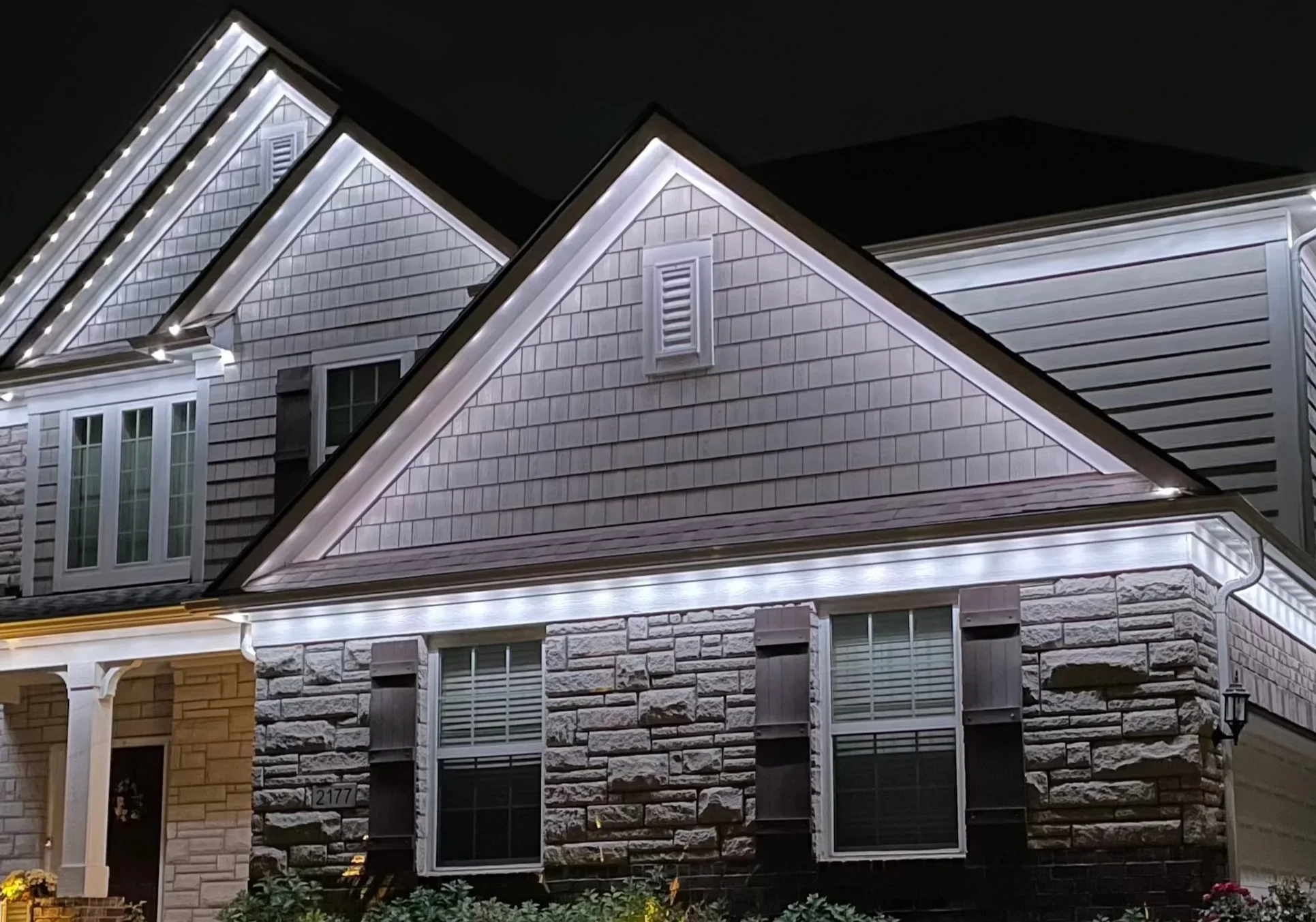 Nighttime view of a house with exterior lighting along the roofline and windows. The house features a combination of stone and siding on its facade, with multiple peaked roofs.