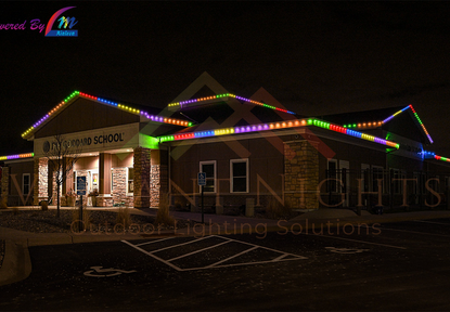 Nighttime exterior view of a school building decorated with colorful LED strip lights along the roofline, with a parking lot in front.