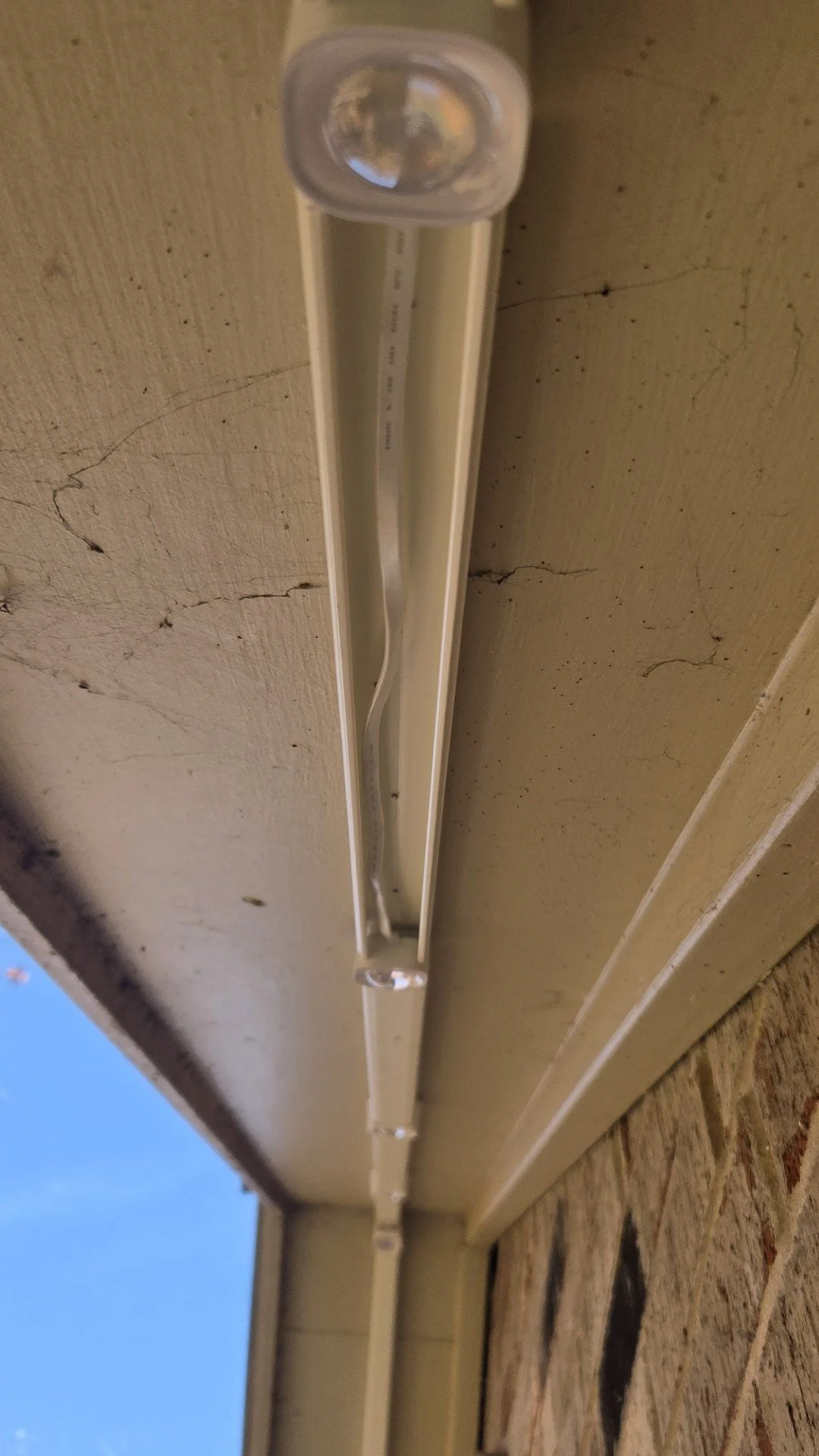 Close-up of a white LED outdoor light fixture attached to a beige porch ceiling next to a brick wall.