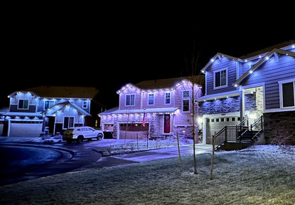 Row of illuminated houses decorated with holiday lights at night, with snow on the ground and parked cars in the scene.