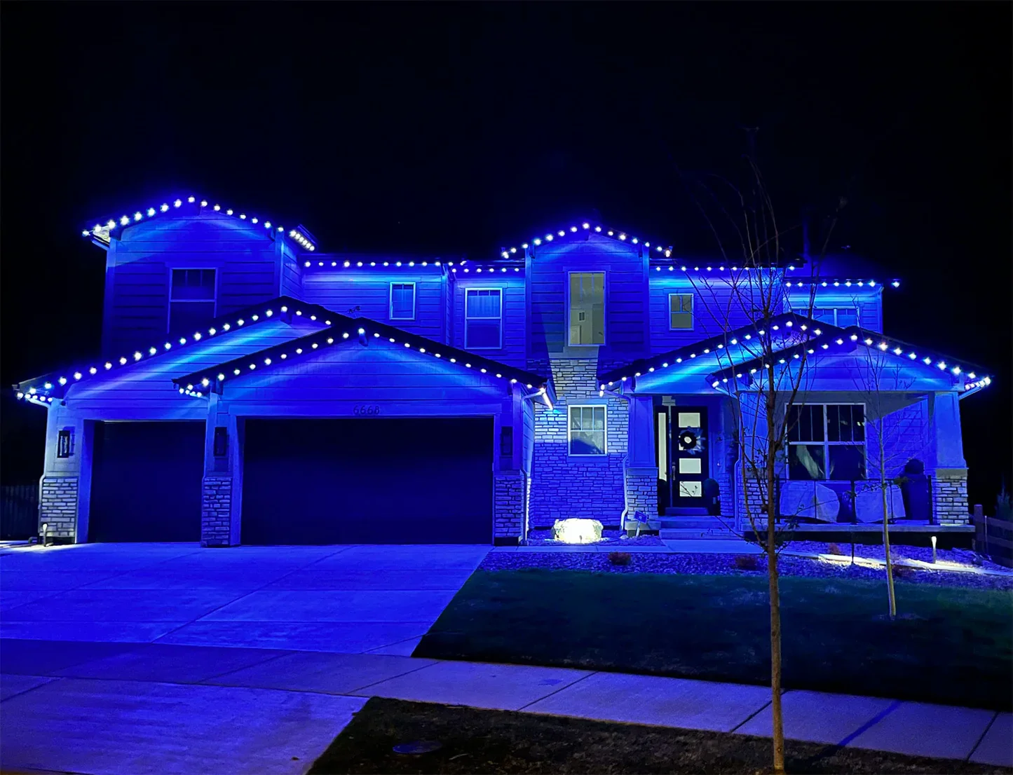 A two-story house decorated with blue Christmas lights at night, with a driveway, small leafless tree, and a front porch with cushions.