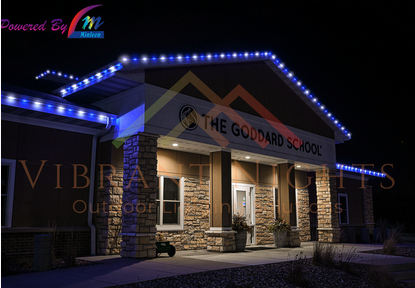Nighttime exterior of The Goddard School with blue and purple Christmas lights outlining the roof, stone pillars at the entrance, and a sign displaying the school name.
