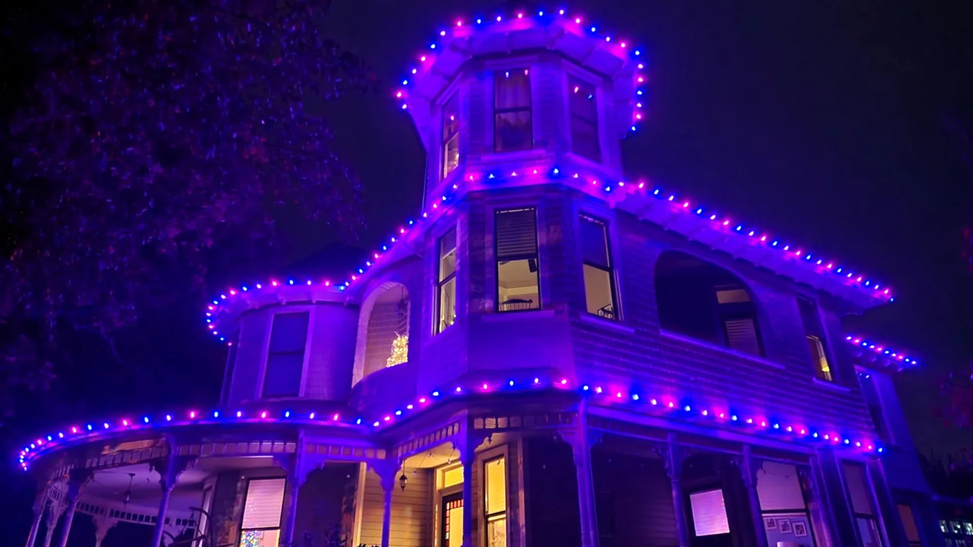 A Victorian-style house decorated with purple and pink string lights on the roofline, windows, and balcony, illuminated at night.
