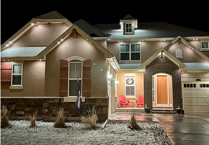Front view of a well-lit house at night with a porch, red chairs, and a pathway with some dry plants in the yard.