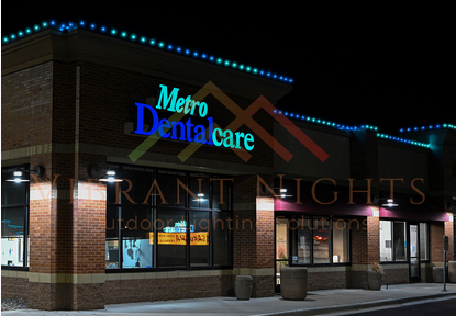 Photo of a Metro Dentalcare building at night with illuminated signage and windows, brick exterior, and decorative blue lights along the roofline.