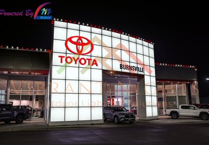 Lighted Toyota dealership building at night with large illuminated signs, including Toyota logo, text reading 'Burnsville,' and dealership vehicles parked outside.