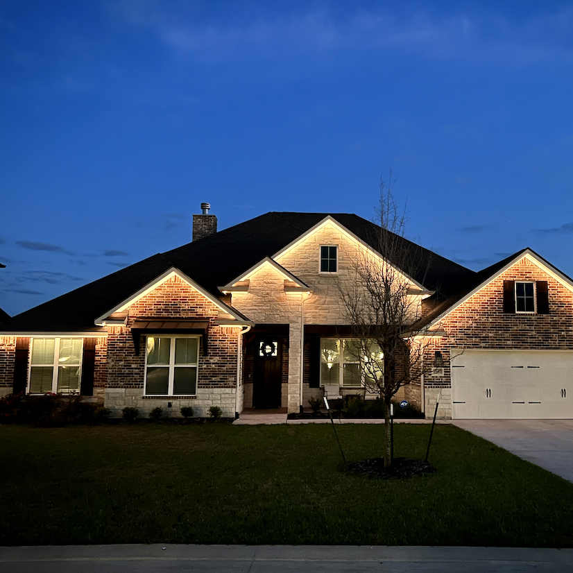 A house with brick and stone exterior, illuminated at dusk, with a front yard and young tree.