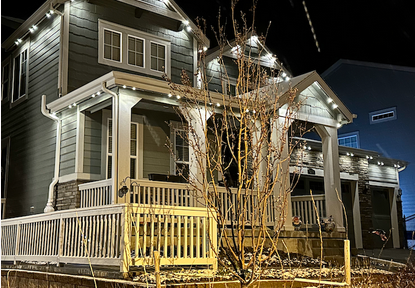 A two-story house decorated with string lights at night, with a wooden front porch, stairs, and a leafless tree in front.
