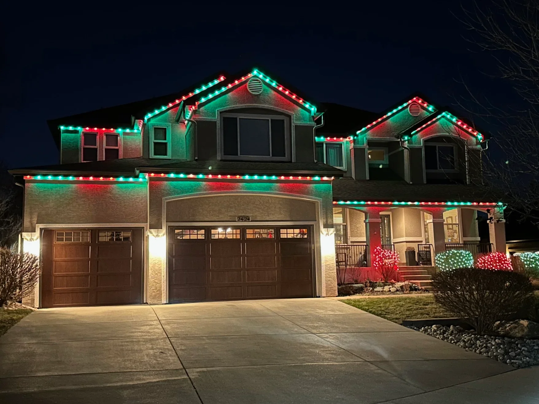 A two-story house decorated with colorful Christmas lights at night, including red and green lights outlining the roof and windows, with some bushes decorated with red and green lights in the front yard.