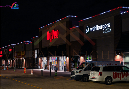 Nighttime view of a shopping center parking lot with Hy-Vee and Wahlburgers stores, cars parked, and colorful string lights on the building roof.