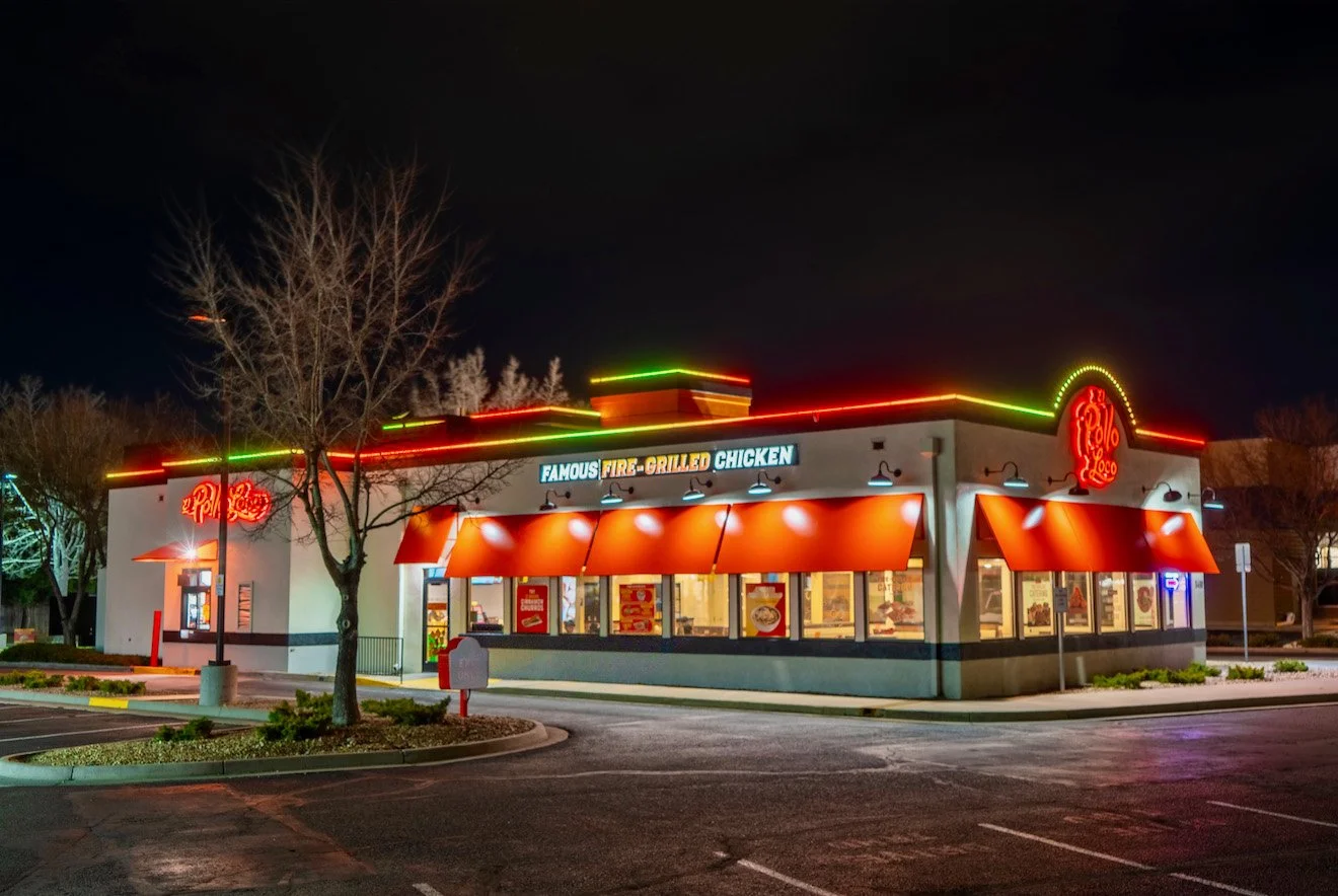 Nighttime view of a fast-food restaurant with neon signs and red awnings, illuminated parking lot, leafless trees surrounding the building.