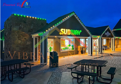 Exterior of a Subway restaurant at dusk with green and yellow signage, outdoor seating, and festive green lighting on the roof.