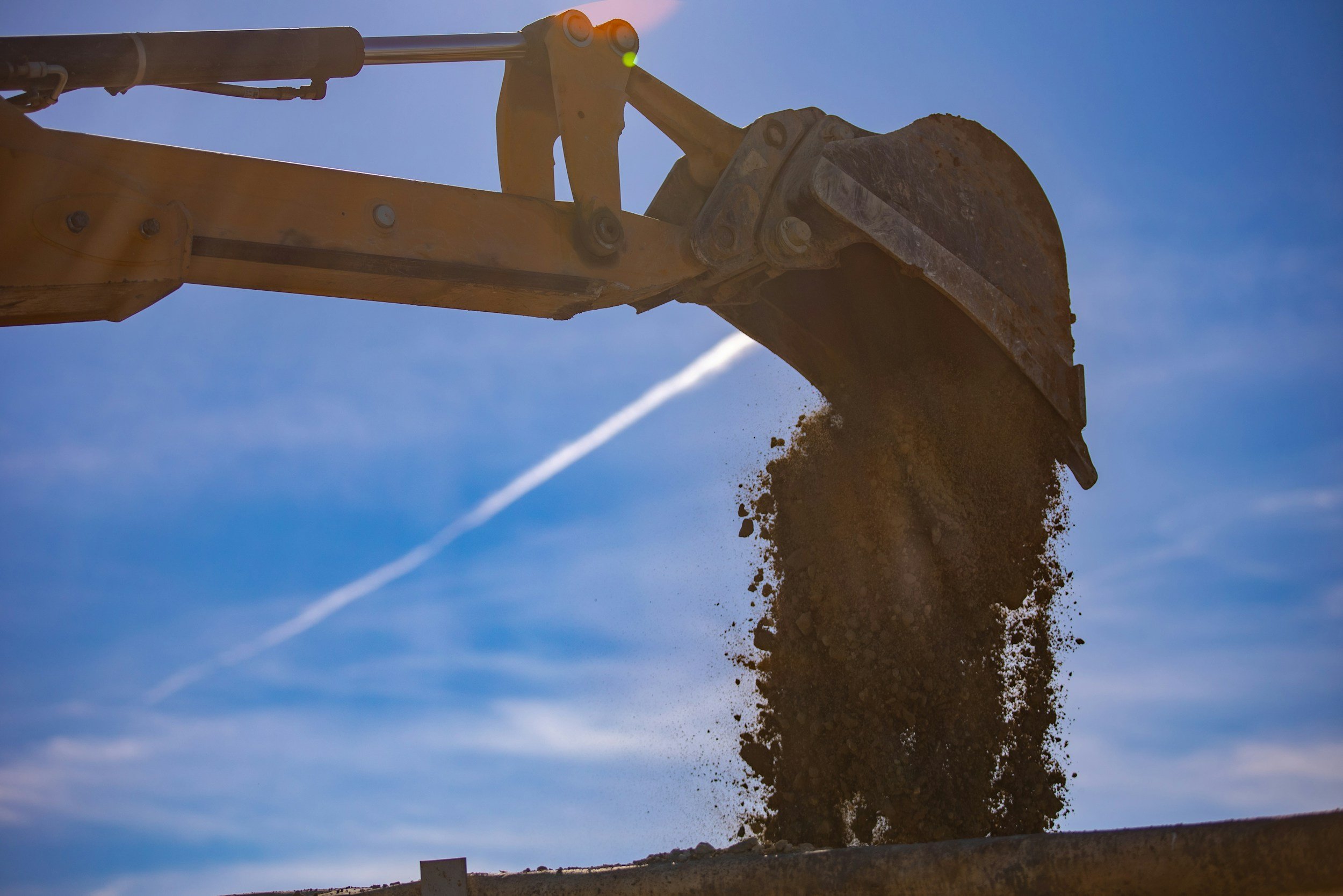 Close-up of a construction excavator bucket dumping soil against a blue sky with wispy clouds.