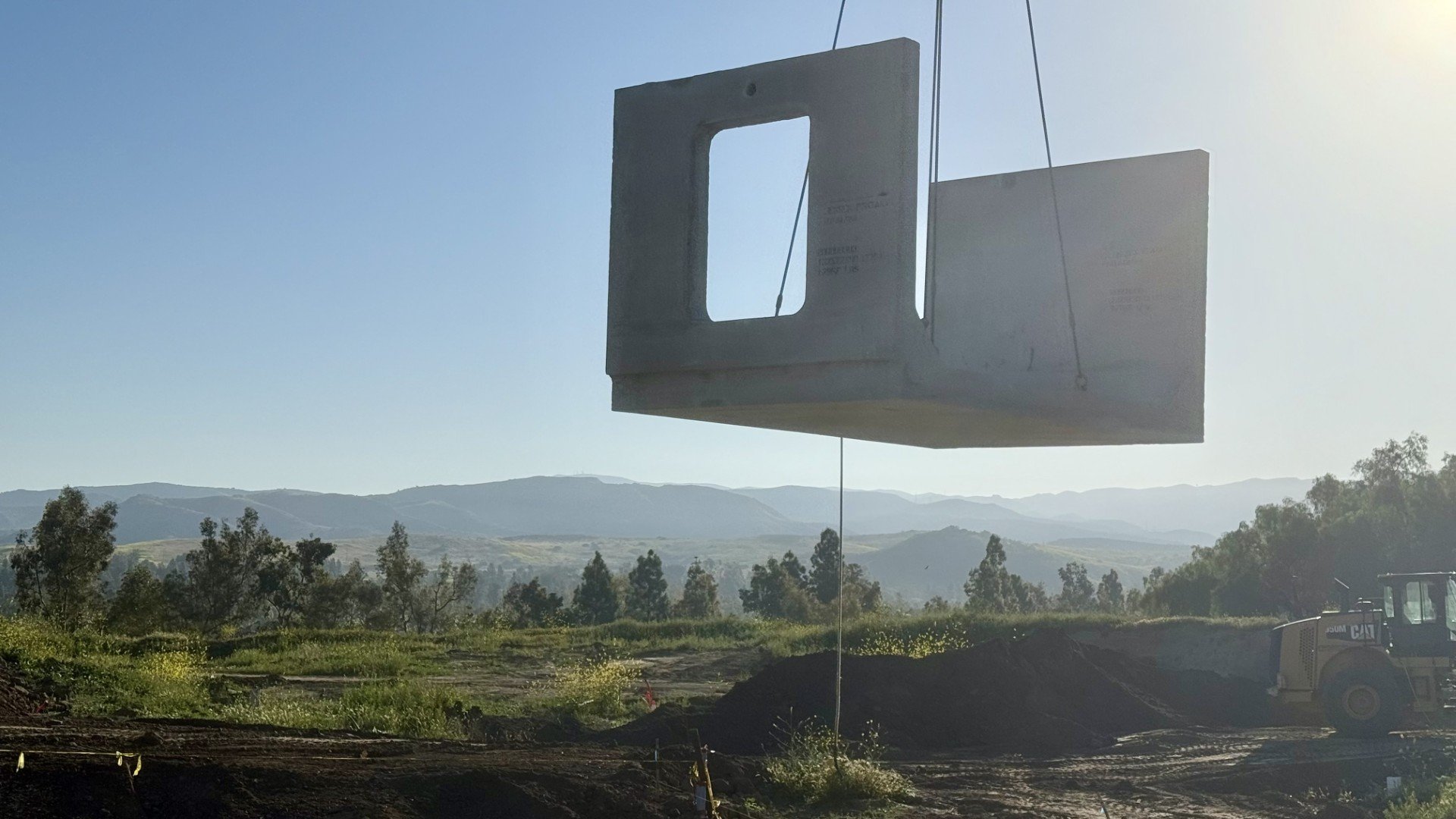 Large concrete prison cell module being lifted by crane at a construction site with hills and trees in the background.