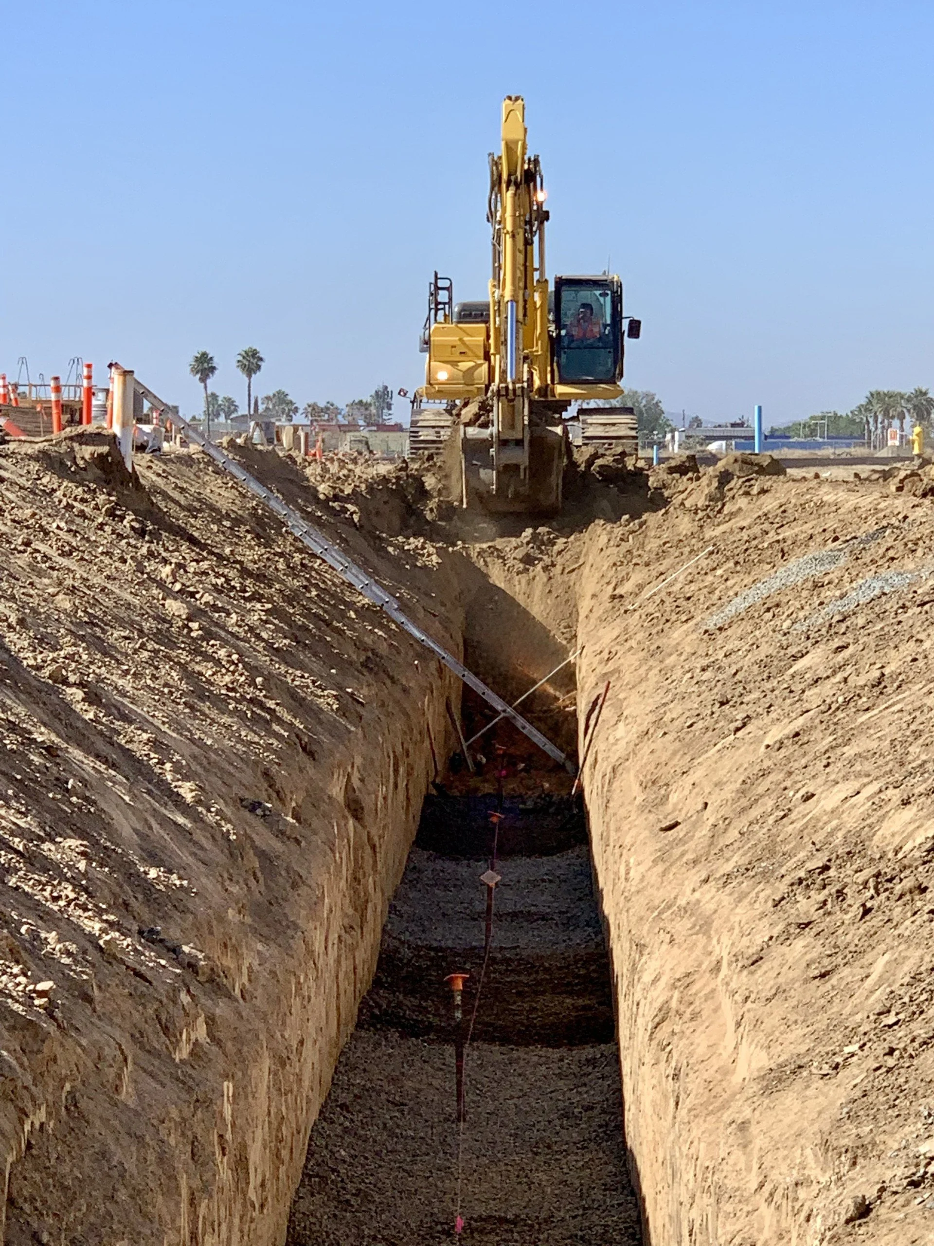 A yellow excavator is working on a construction site, digging a deep trench in the dirt under a clear blue sky.