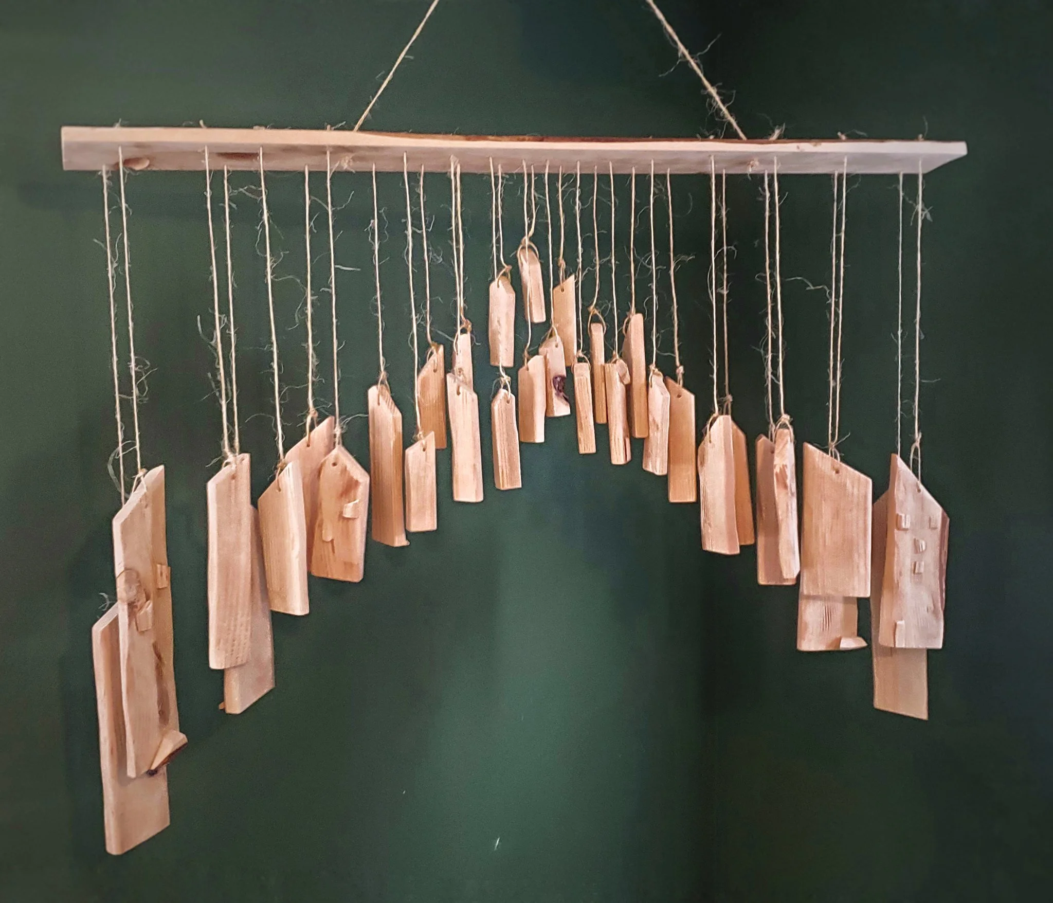 A handmade wooden advent calendar with small tags hanging from strings, displayed on a green wall.