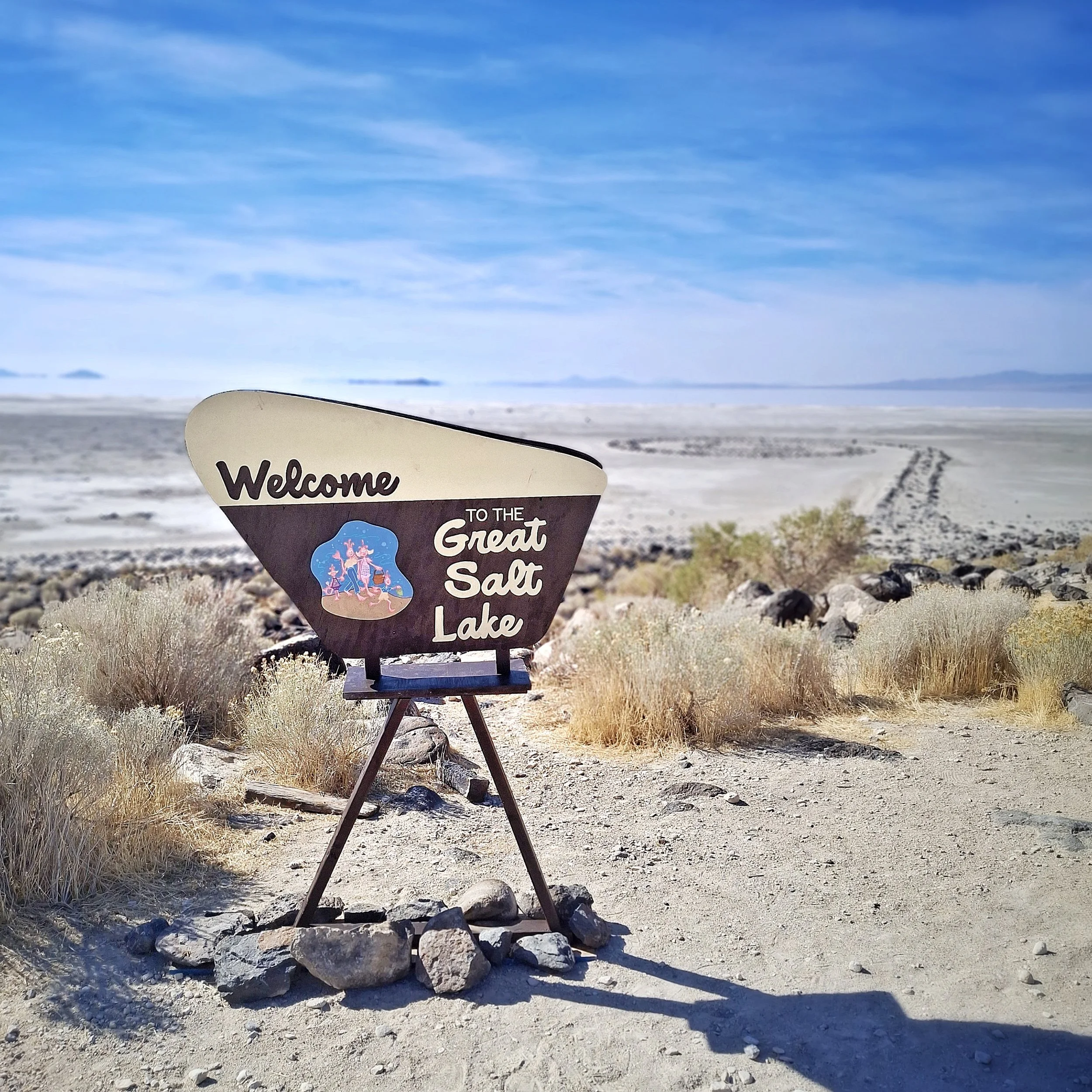 Sign welcoming visitors to the Great Salt Lake, with a desert landscape, rocks, and blue sky in the background.