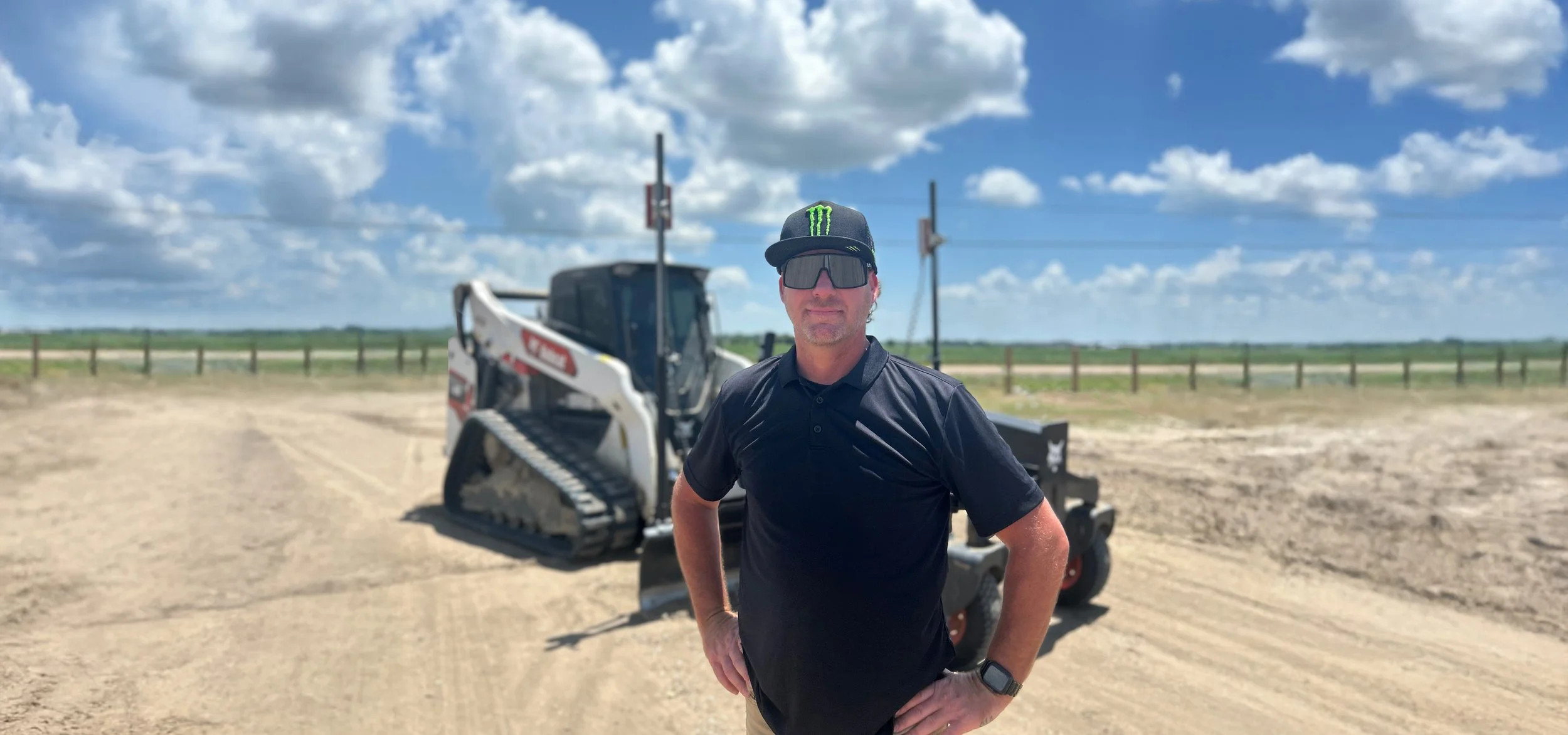 Chris Sandifer, Owner and Operator, with landscaping equipment on a dirt lot with a clear blue sky and puffy clouds.