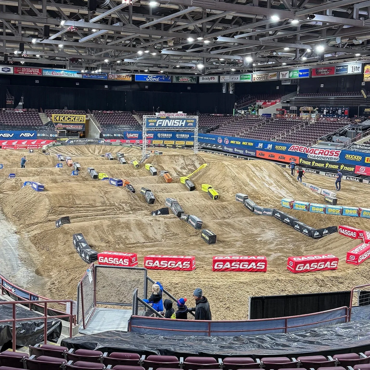 Arenacross track with jumps and obstacles, surrounded by seating and banners, with a few spectators and officials preparing for a race.