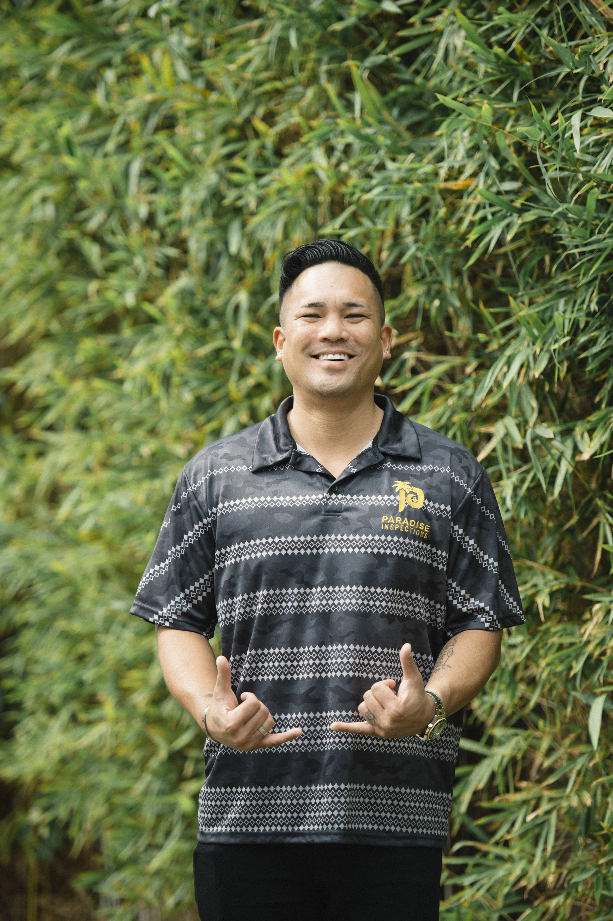 A man smiling and making thumbs-up gestures outdoors, standing in front of greenery, wearing a black patterned polo shirt with a logo that reads "Paradise Inspections".