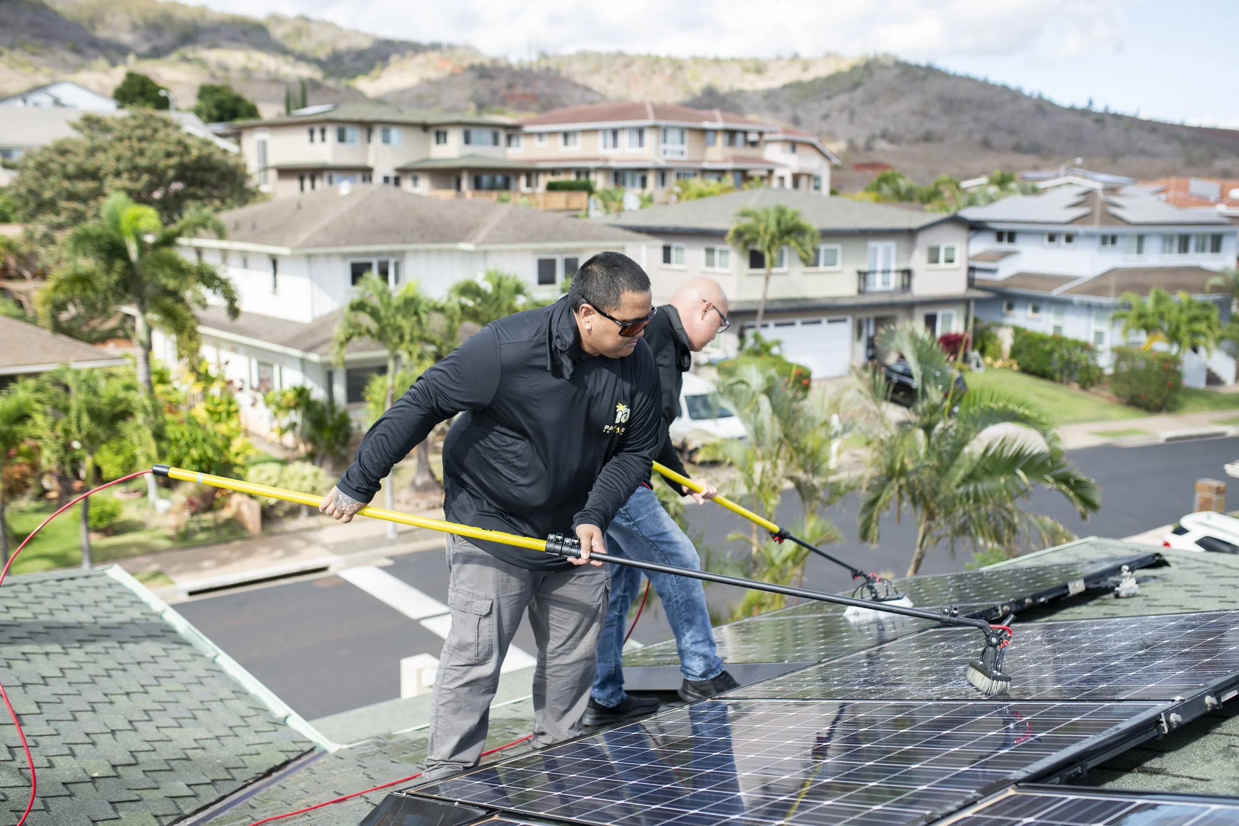Two men are installing or cleaning solar panels on a house roof during the daytime, with a suburban neighborhood and hills in the background.