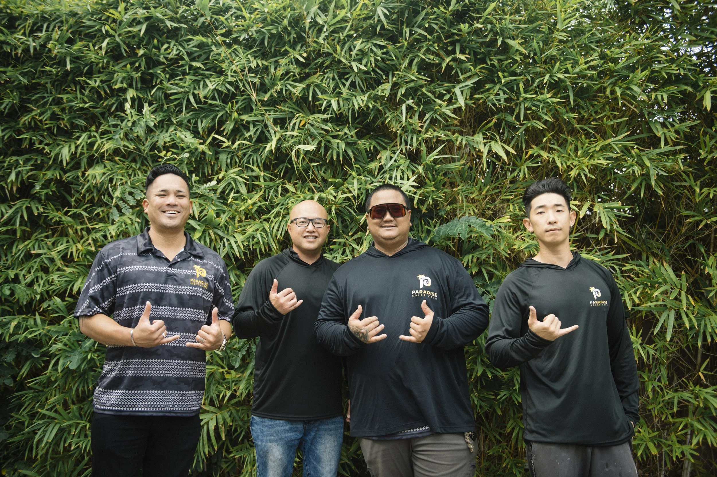 Group of four men standing outdoors in front of leafy green foliage, posing for the camera with hand signs and smiling.