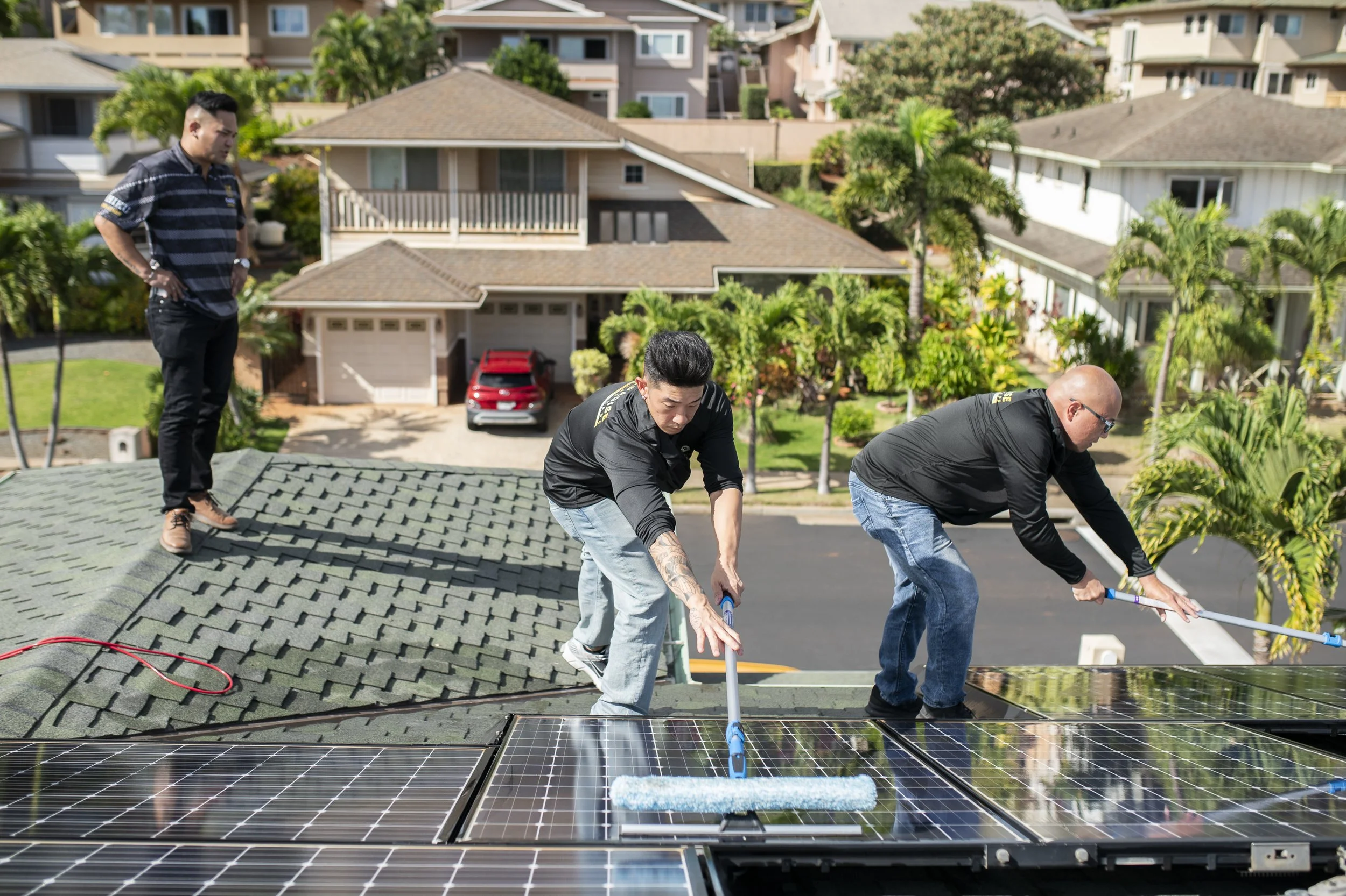 Three men install solar panels on a rooftop in a residential neighborhood, with houses, trees, and a street visible in the background.
