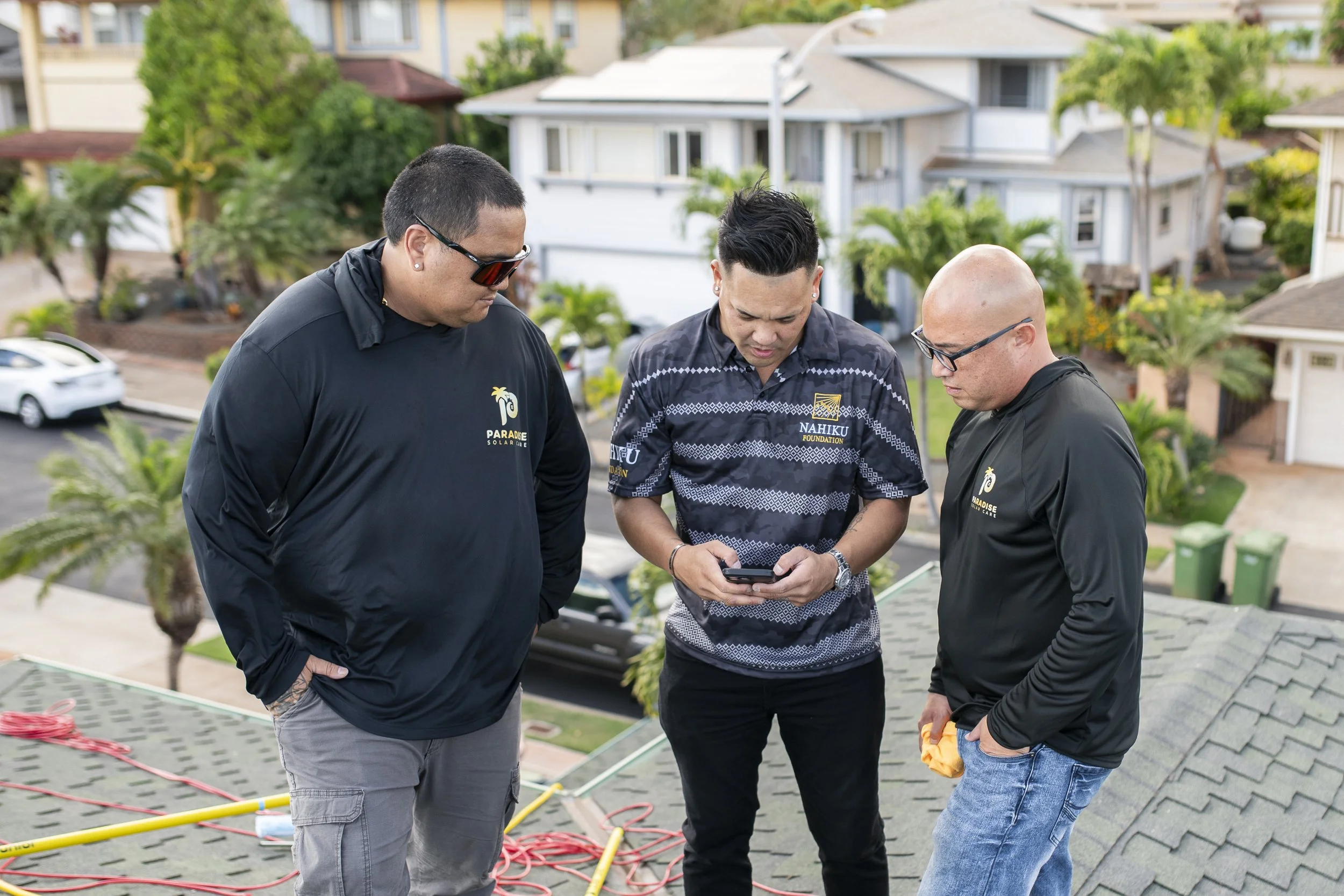 Three men standing on a rooftop looking at a smartphone, with houses and trees in the background.
