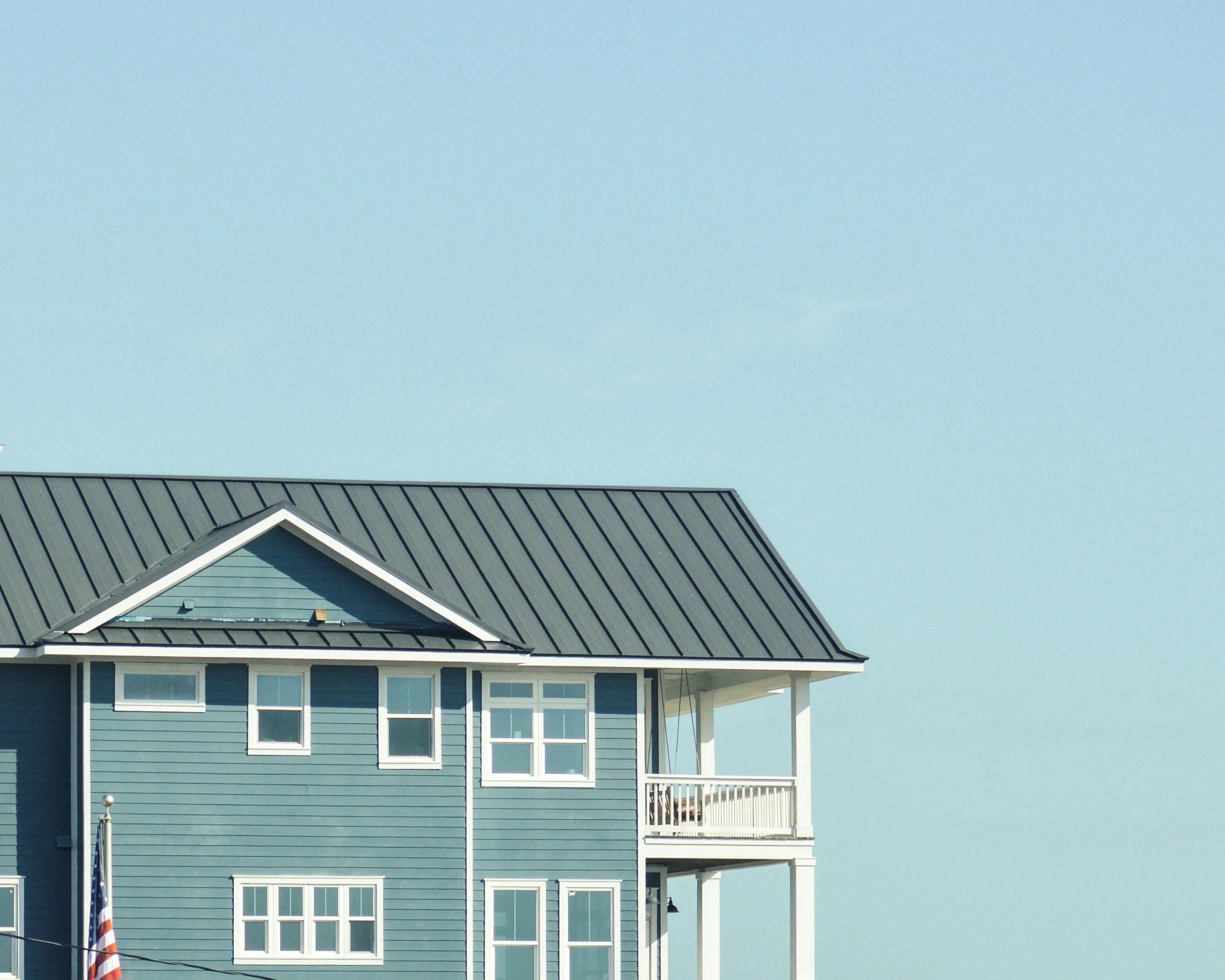 A blue multi-story house with white trim and a metal roof, featuring a small balcony and multiple windows, under a clear blue sky.