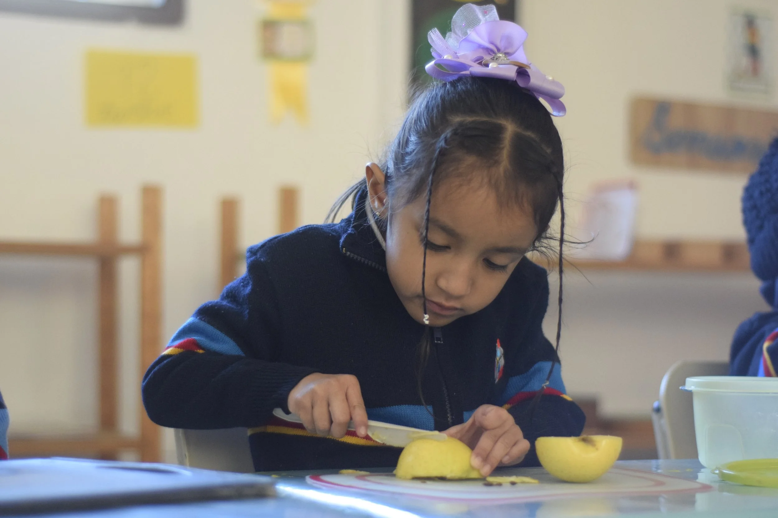 Niña cortando una papa en una mesa de aula escolar.