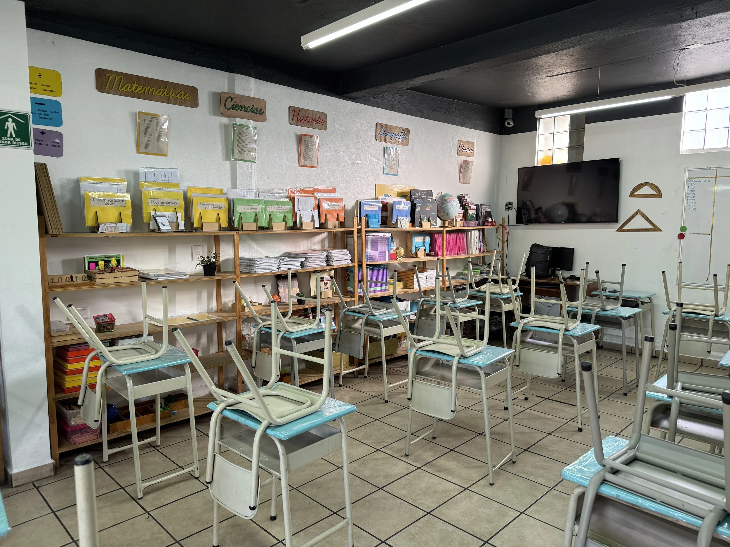 Aula escolar con sillas apiladas, estanterías con libros y materiales escolares, cuadros con etiquetado de asignaturas como Matemáticas, Ciencias, Historia, Literatura y Física, en una pared, y televisión en la esquina.