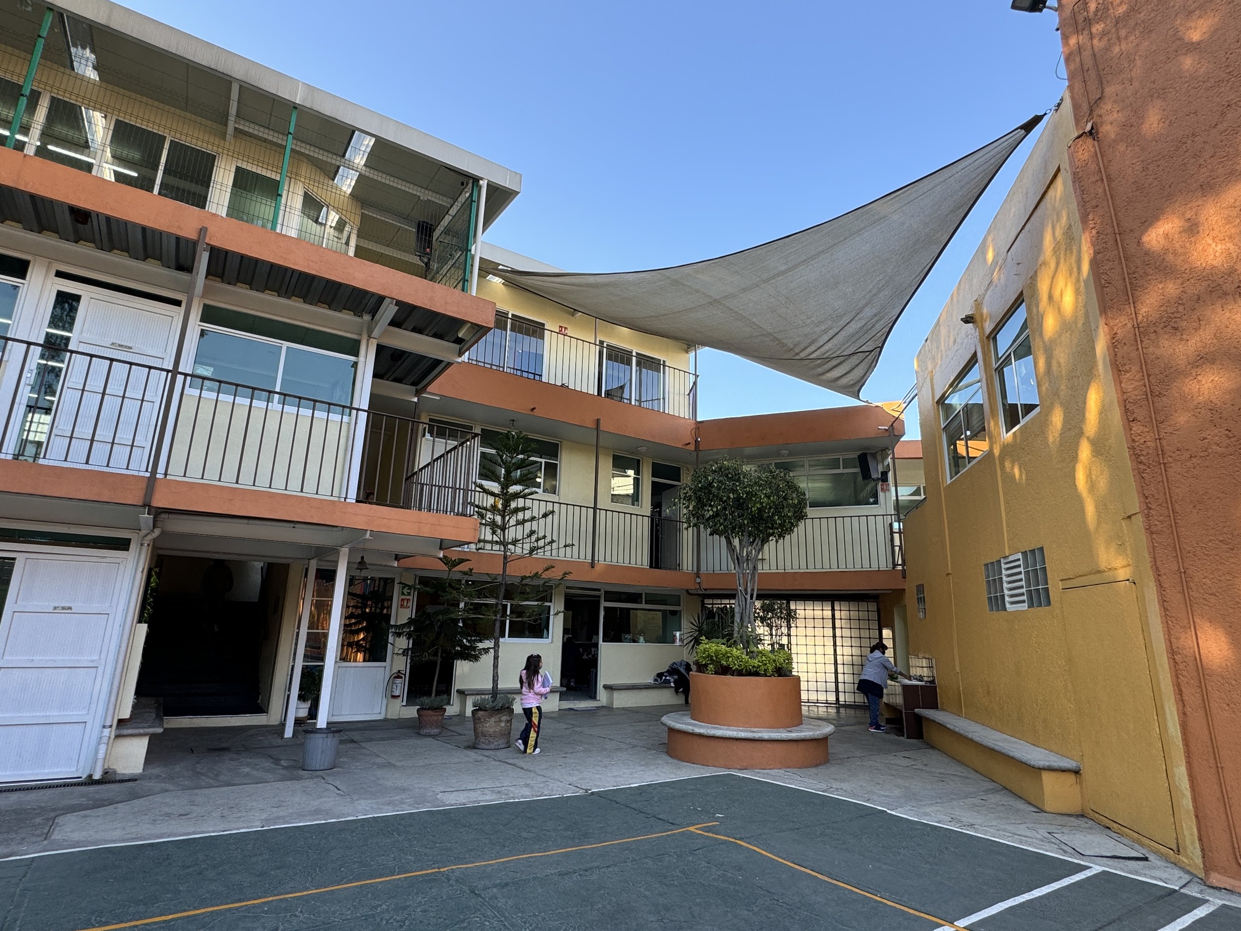 Vista de un patio interior de edificio con niños jugando y una cancha, con paredes amarillas y naranjas, una planta en maceta grande y toldos de sombra.