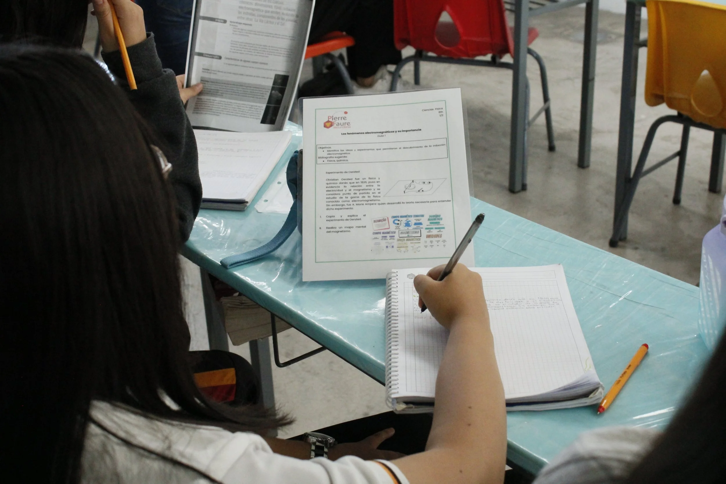 Estudiantes en un aula tomando apuntes, con materiales escolares y una hoja con información sobre fenómenos electromagnéticos en la mesa.