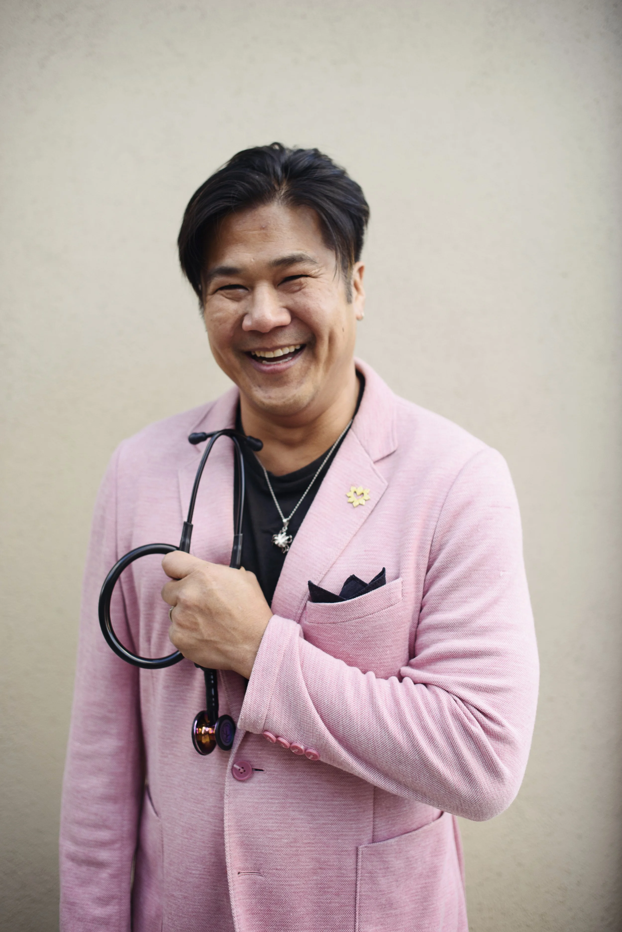 A man smiling and holding a stethoscope, wearing a pink blazer, black shirt, and layered necklaces, standing against a beige wall.
