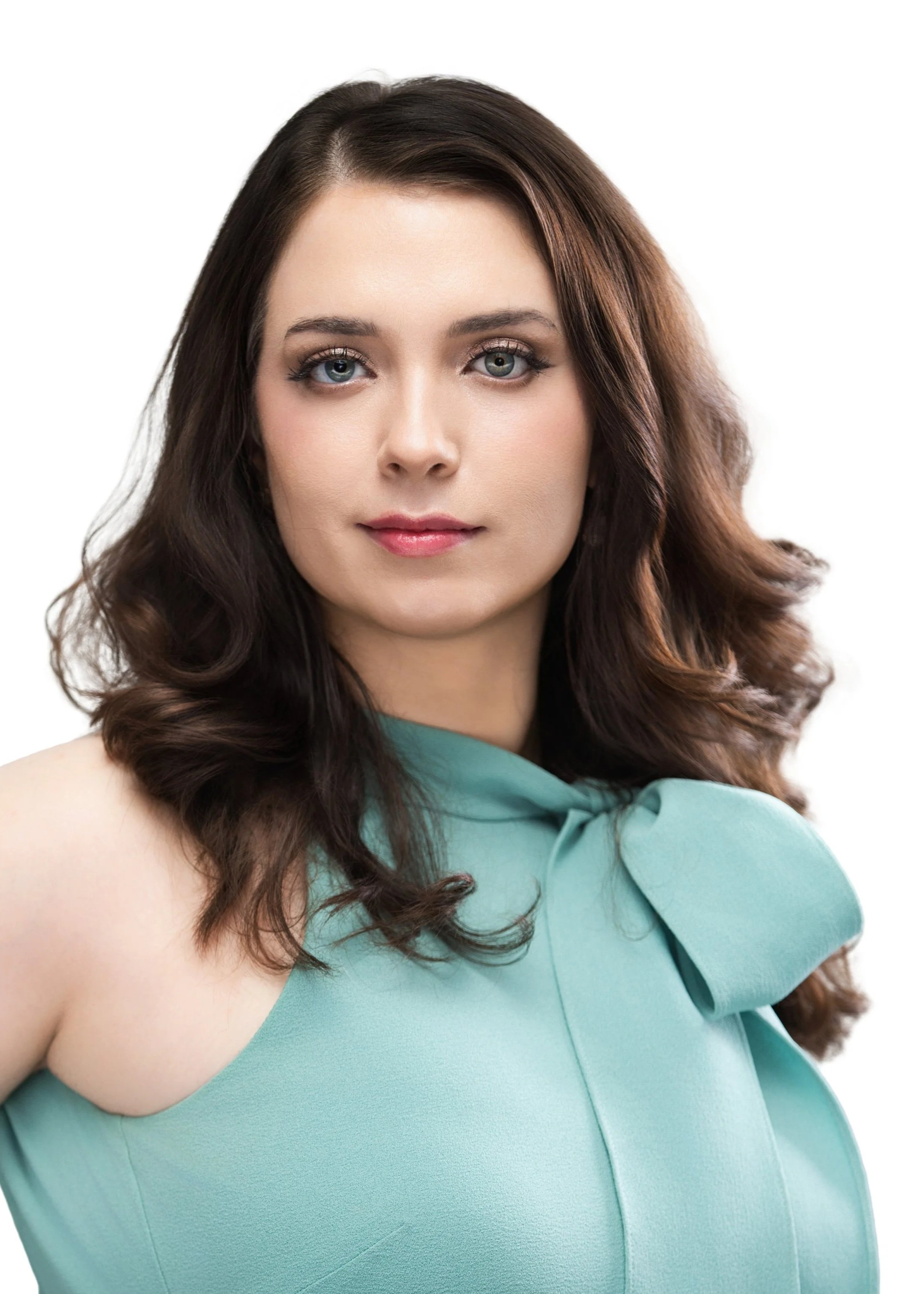 A woman with long, wavy brown hair and blue eyes, wearing a light blue sleeveless top with a bow detail on the shoulder, against a plain white background.