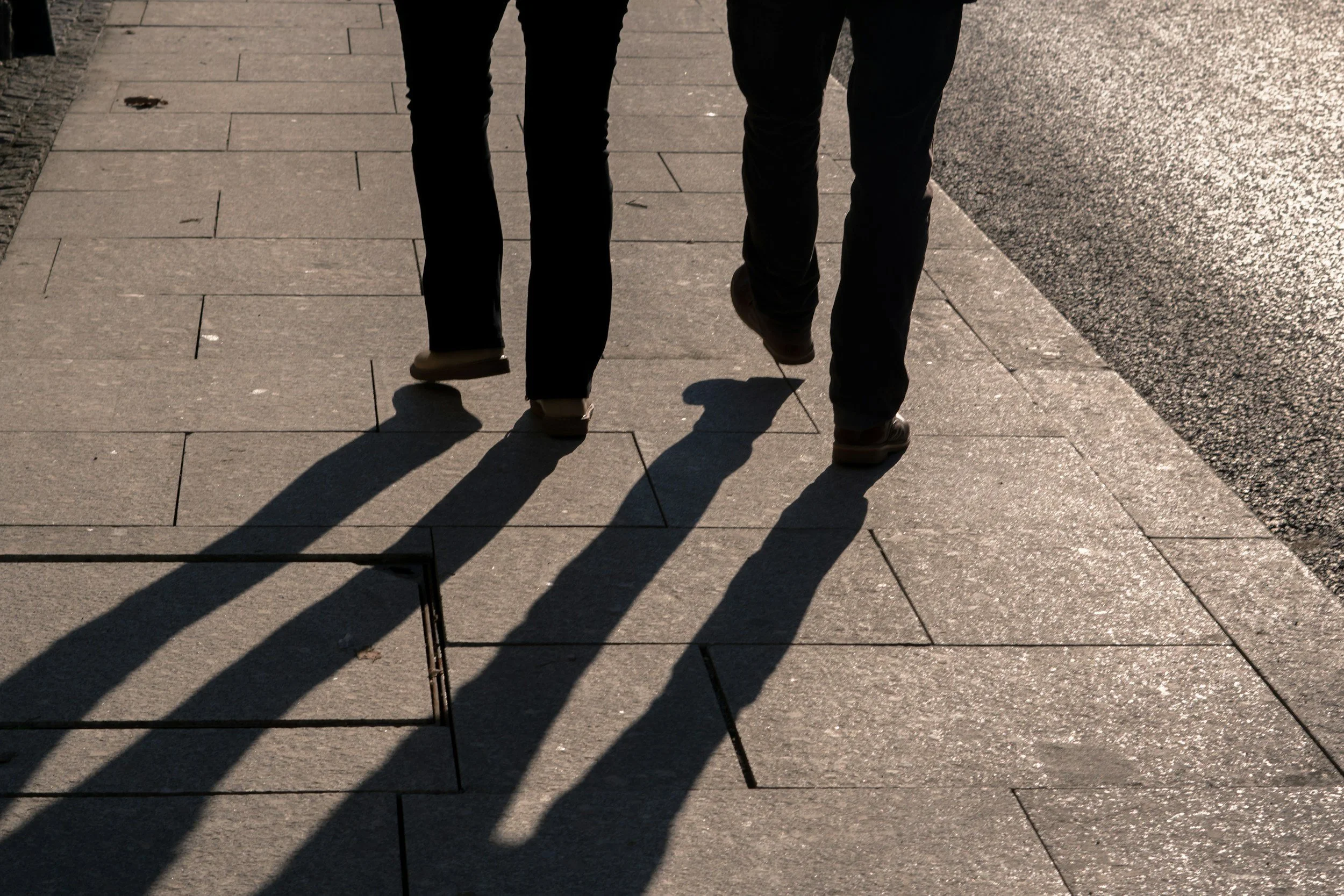 Two people walking on a sidewalk with shadows cast in the sunlight.
