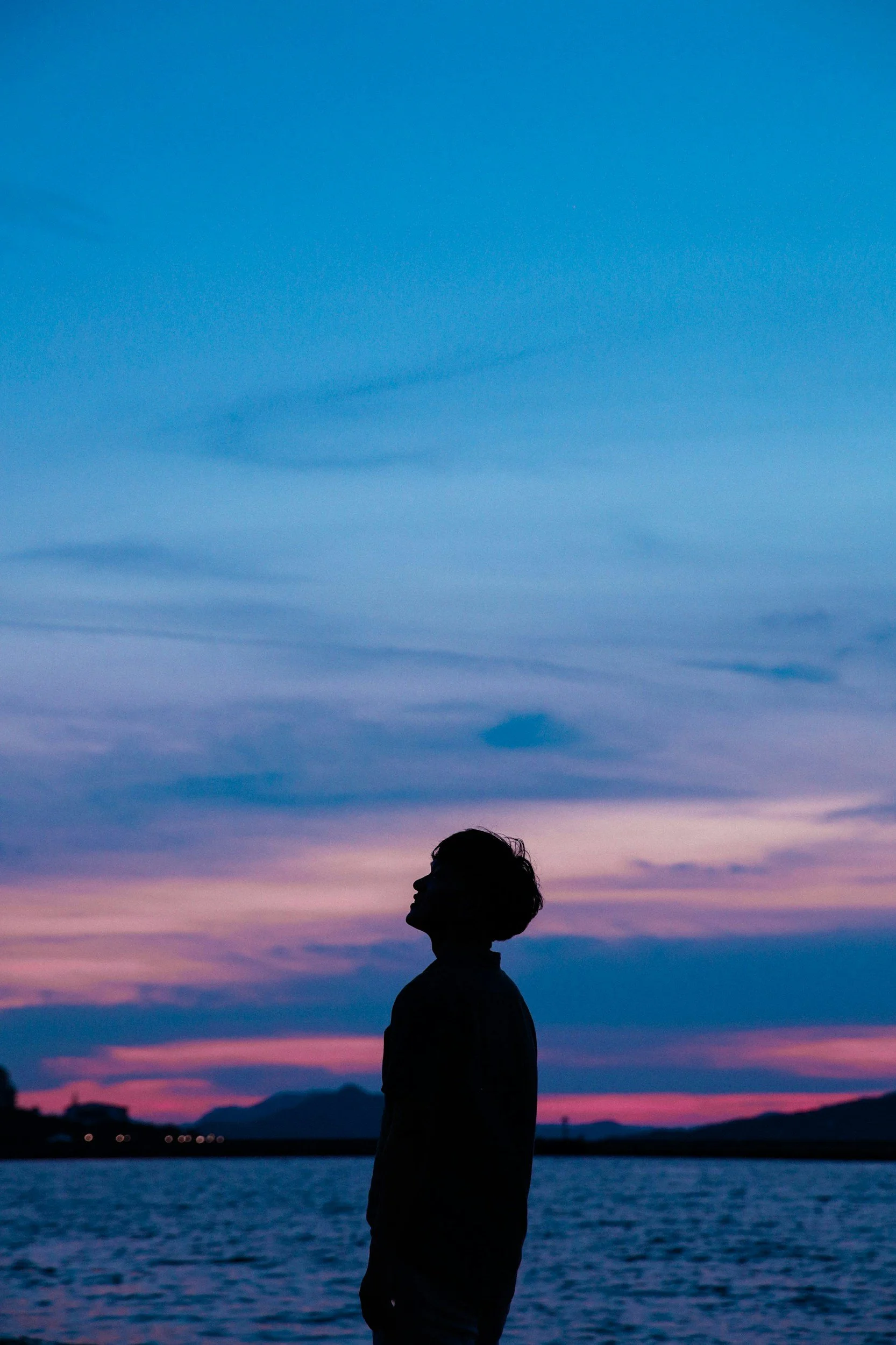 Silhouette of a person standing near a body of water during sunset or dusk, with colorful sky and mountains in the background.